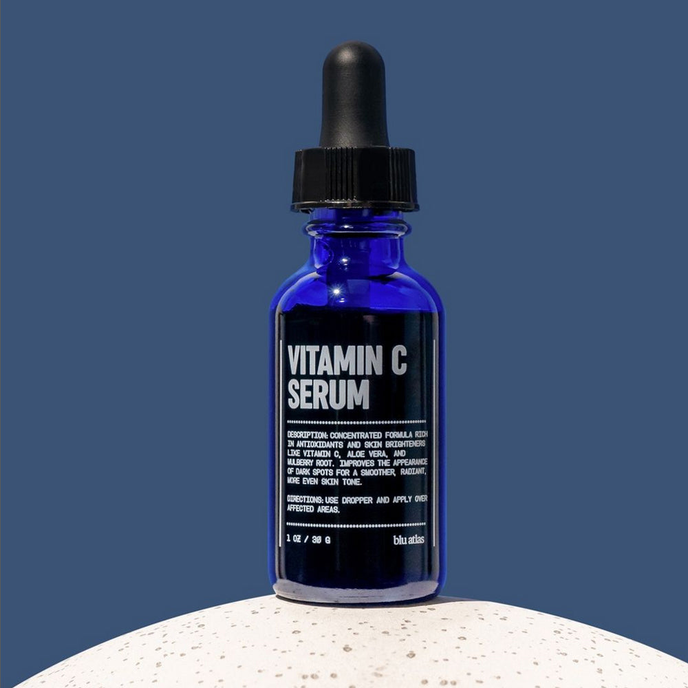 A blue glass dropper bottle of Blu Atlas Vitamin C Serum stands upright on a white textured surface against a plain blue background, showcasing its brightening ascorbic acid formula.