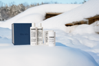The Blu Atlas Body Care Essentials Set, featuring bottles of shampoo, conditioner, and deodorant in a navy box, is displayed outdoors on snow with wooden structures and trees in the background.