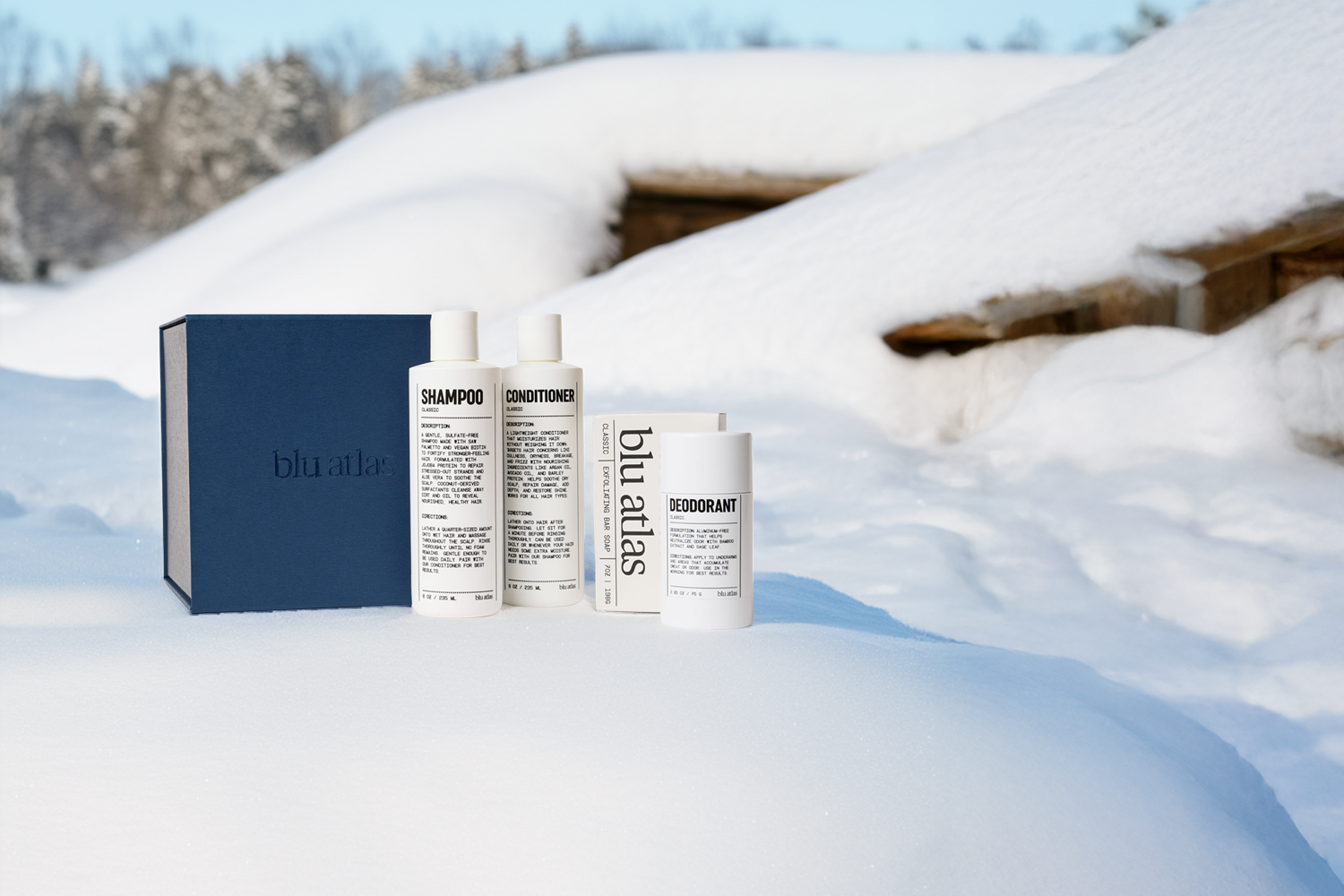 The Blu Atlas Body Care Essentials Set, featuring bottles of shampoo, conditioner, and deodorant in a navy box, is displayed outdoors on snow with wooden structures and trees in the background.