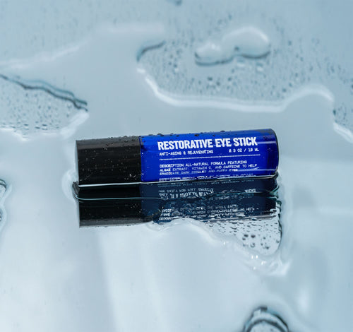 A sleek blue tube labeled Blu Atlas Eye Stick with a black cap rests on a wet, reflective surface surrounded by water droplets, emphasizing its caffeine roll-on formula for reducing puffiness and dark circles.