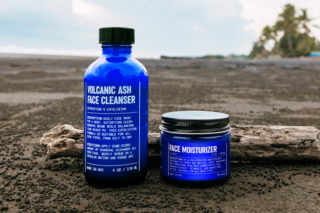 Two blue Blu Atlas Face Essentials containers rest on driftwood at the beach, with ocean waves and palm trees behind them—perfect for tackling early signs of aging.