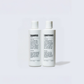 Two white Blu Atlas Hair Essentials bottles (Shampoo + Conditioner) stand side by side, each labeled in black text—vegan biotin formulas ideal for daily hair care.