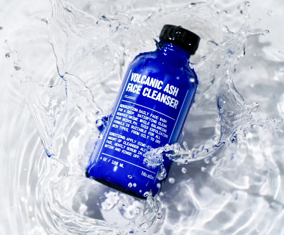 Close-up of a blue bottle labeled Volcanic Ash Face Cleanser with water splashes surrounding it.