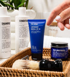 A hand reaches for a Blu Atlas blue tube of Lightweight Mineral SPF 30 with UVA/UVB protection in a basket, surrounded by shampoo, conditioner, face moisturizer, and a shaving brush on the bathroom counter.