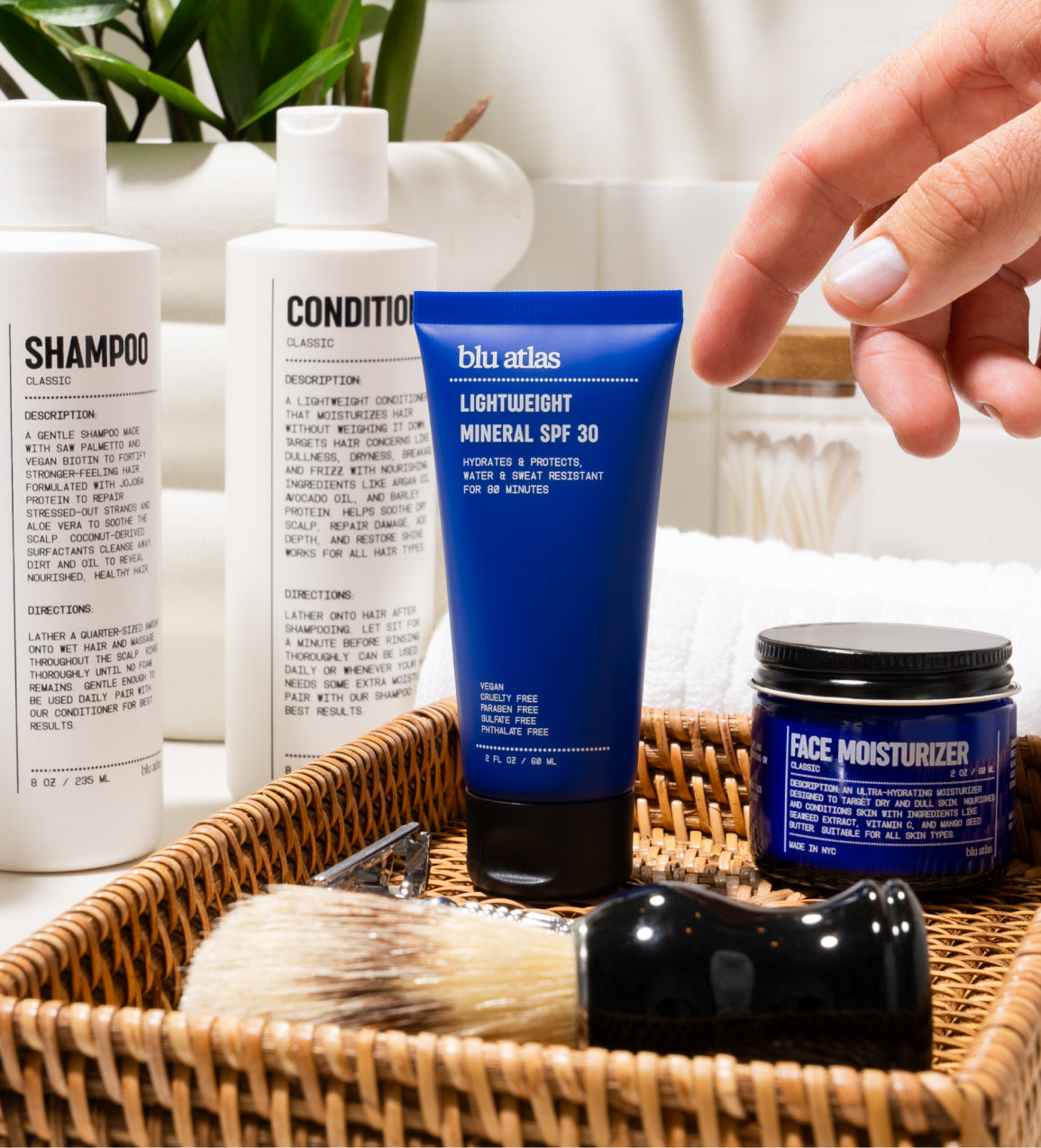 A hand reaches for a Blu Atlas blue tube of Lightweight Mineral SPF 30 with UVA/UVB protection in a basket, surrounded by shampoo, conditioner, face moisturizer, and a shaving brush on the bathroom counter.