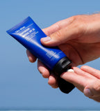 A person applying sunscreen from a blue tube labeled Protect & Moisturize Duo by Blu Atlas onto their finger against a clear sky, benefiting from natural skincare with UVA and UVB protection, ensuring water- and sweat-resistant coverage.