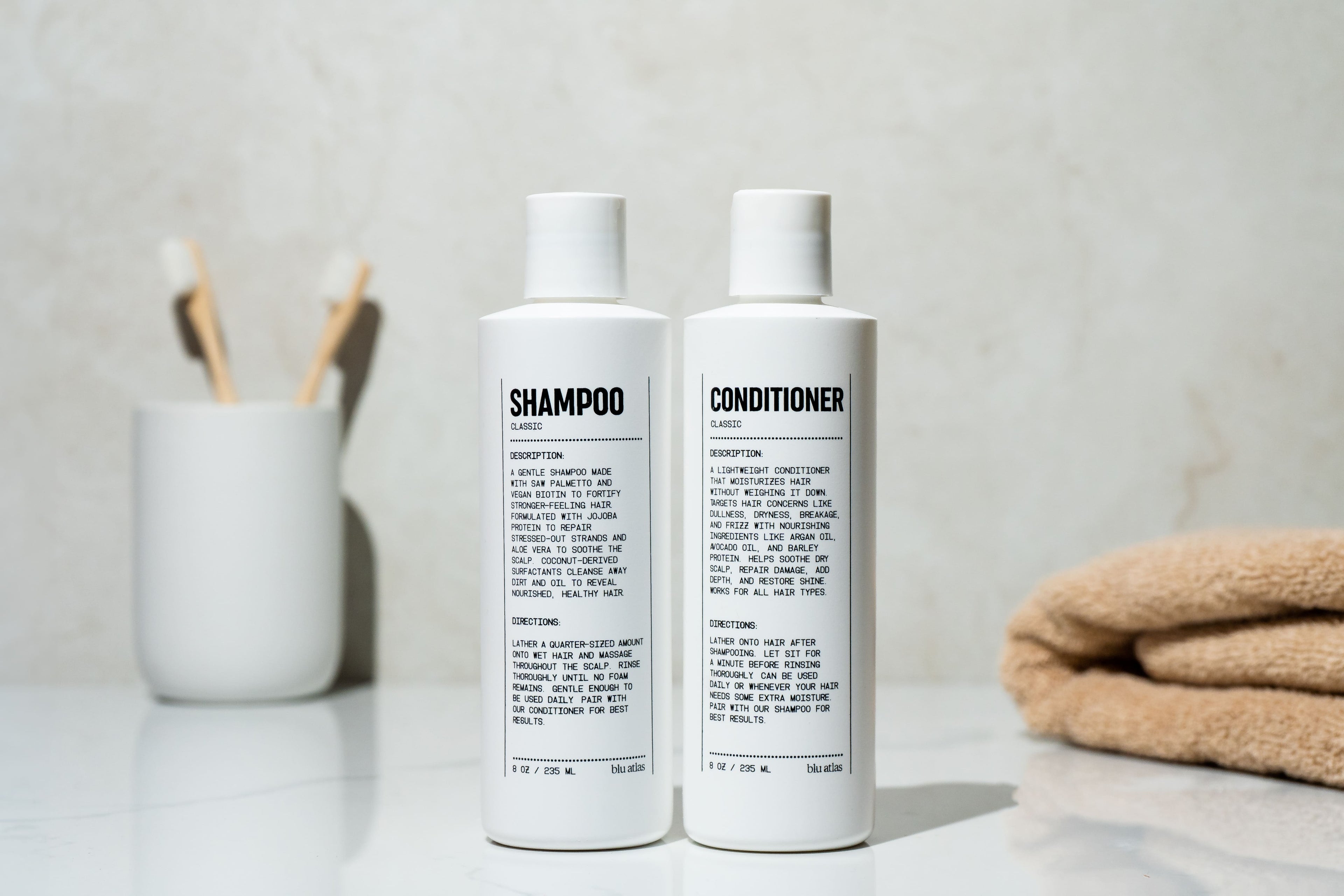 Two white Blu Atlas Hair Essentials bottles (Shampoo + Conditioner) rest on the bathroom counter, ideal for daily hair care. A cup of toothbrushes and a folded towel complete this clean vegan routine.