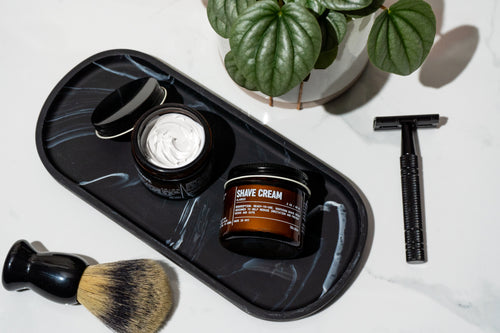 Shaving products including a jar of shave cream, brush, and razor on a decorative tray with a plant in the background.