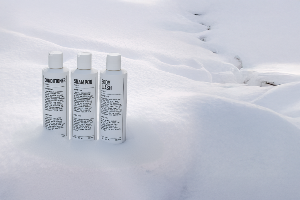 Three white Blu Atlas Shower Bundle bottles labeled conditioner, shampoo, and body wash stand upright on snow, their black text visible—a great grooming set for men seeking natural care.