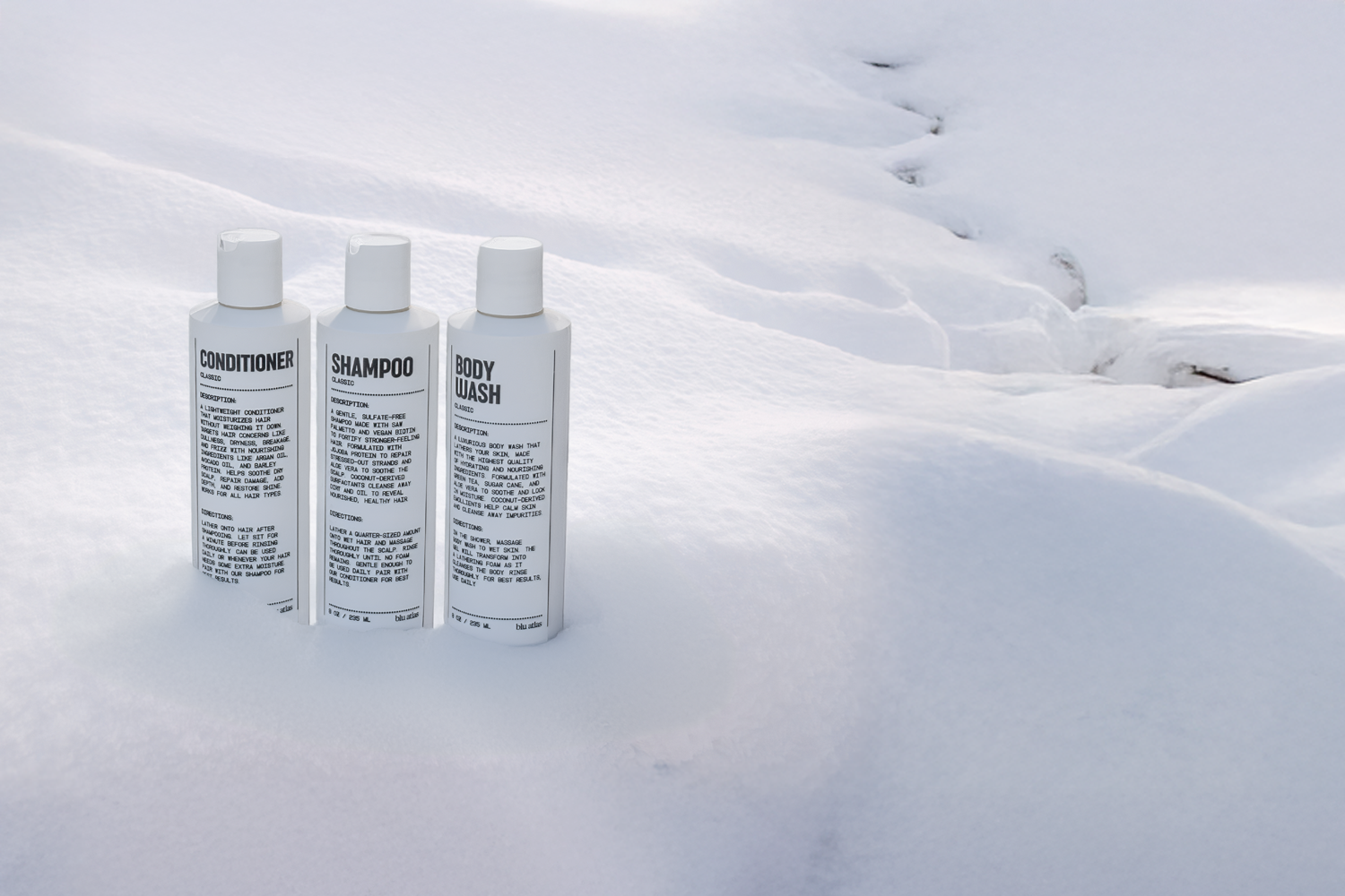 Three white Blu Atlas Shower Bundle bottles labeled conditioner, shampoo, and body wash stand upright on snow, their black text visible—a great grooming set for men seeking natural care.