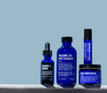 Four blue skincare product containers labeled vitamin C serum, volcanic ash face cleanser, restorative eye stick, and face moisturizer are arranged in a row against a blue background. The Blu Atlas Restorative Eye Stick is featured for dark circle treatment, working like a caffeine eye roll-on to refresh and brighten the under-eye area.