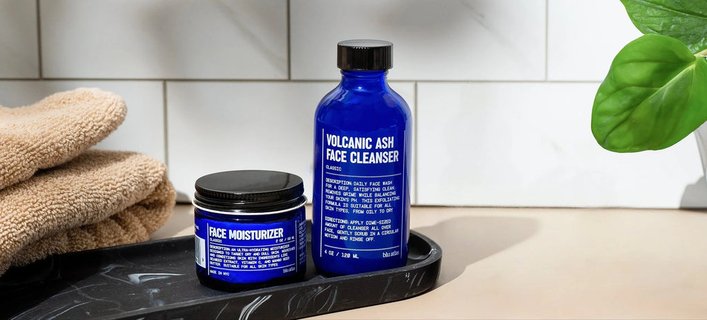 Two blue skincare products on a tray with a towel and plant in the background.