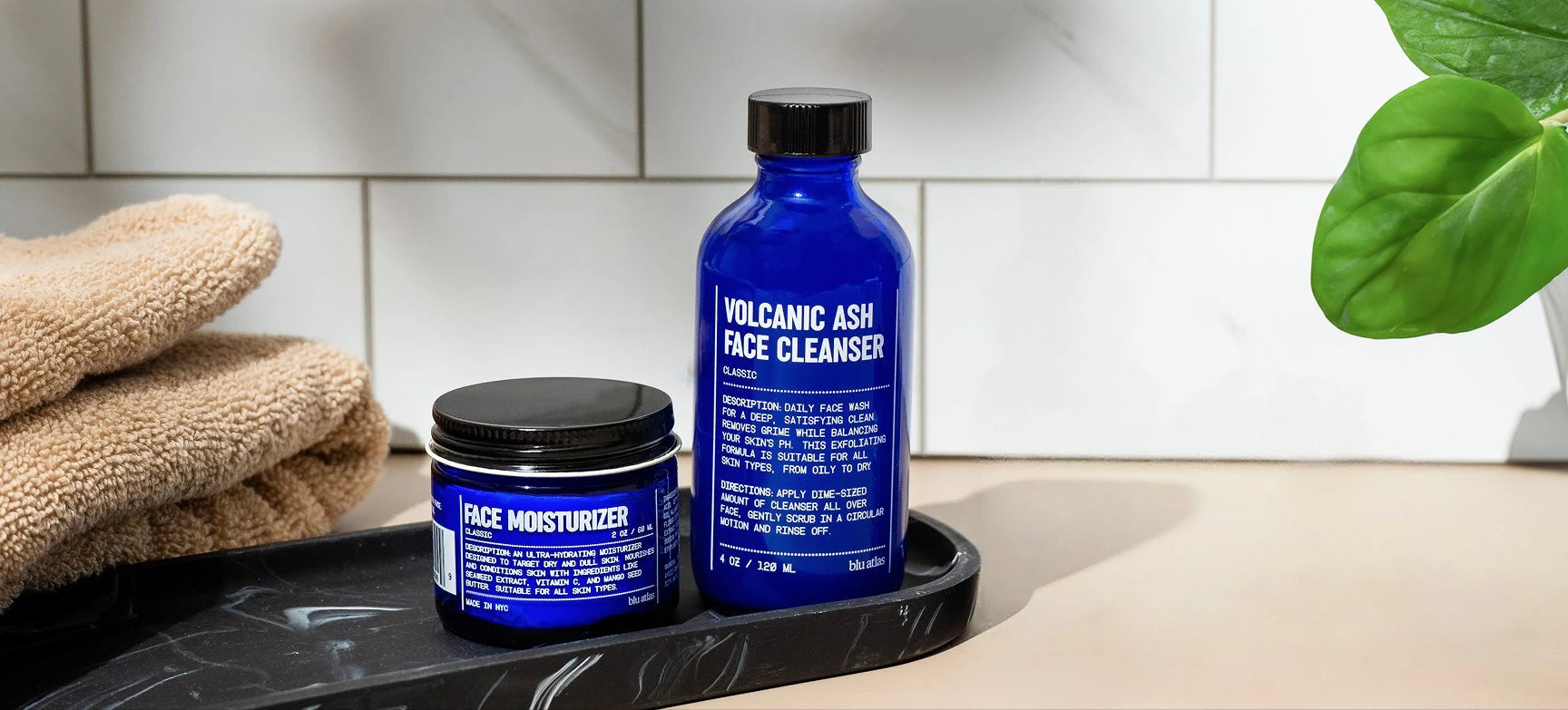 Two blue skincare products on a tray with a towel and plant in the background.