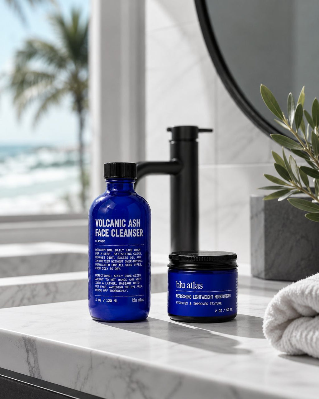 Two blue bottles from Blu Atlas's Summer Face Essentials set rest on a bathroom counter, providing the perfect routine for oily skin. A towel, a plant, and a tranquil beach view complete the refreshing background scene.