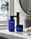 Two blue bottles from Blu Atlas's Summer Face Essentials set rest on a bathroom counter, providing the perfect routine for oily skin. A towel, a plant, and a tranquil beach view complete the refreshing background scene.