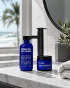 Two blue bottles from Blu Atlas's Summer Face Essentials set rest on a bathroom counter, providing the perfect routine for oily skin. A towel, a plant, and a tranquil beach view complete the refreshing background scene.
