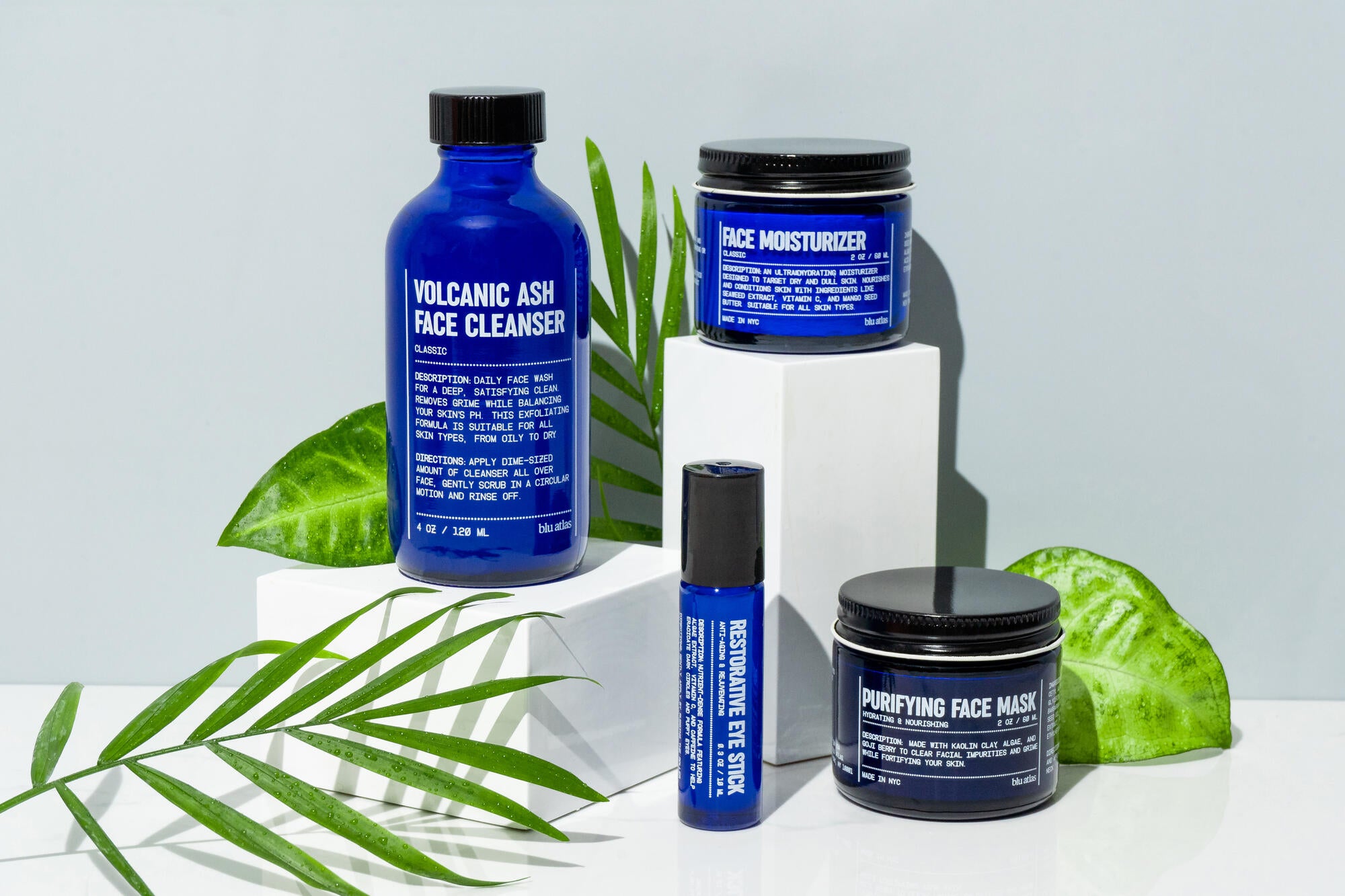 Four blue Blu Atlas skincare containers—exfoliating face wash, moisturizer, eye stick, and Volcanic Ash Face Cleanser—sit on white blocks with green leaves. All products are fragrance free for gentle care.