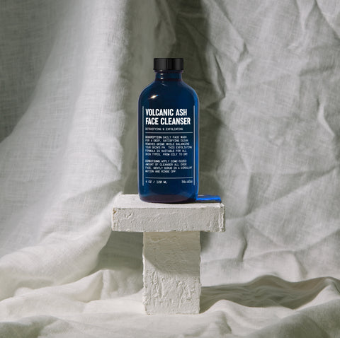 Dark blue bottle of Volcanic Ash Face Cleanser on a small white pedestal with a textured fabric background.