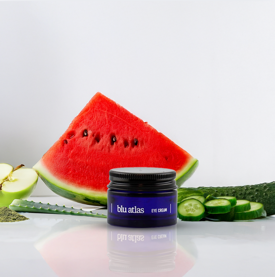 A jar of Blu Atlas Brightening Eye Cream, featuring sodium hyaluronate, is showcased before watermelon, apple, cucumber, and aloe on a white backdrop—ideal for diminishing dark circles and refreshing your skin.