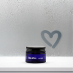 A blue jar of Blu Atlas Brightening Eye Cream rests on a white shelf, offering skin repair. A heart shape is drawn on the foggy glass background, symbolizing its care for dark circles.