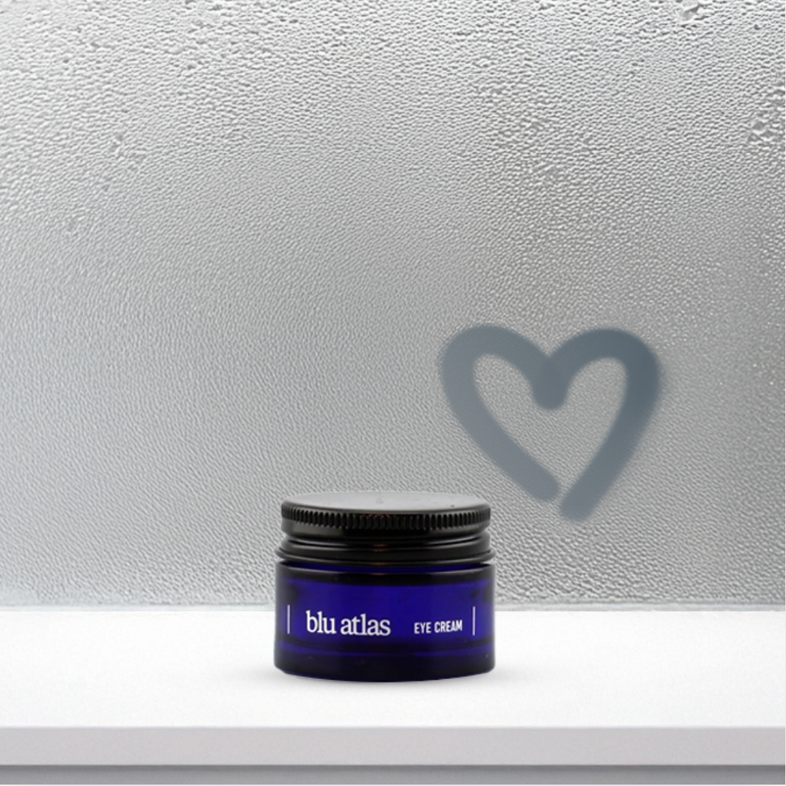 A blue jar of Blu Atlas Brightening Eye Cream rests on a white shelf, offering skin repair. A heart shape is drawn on the foggy glass background, symbolizing its care for dark circles.