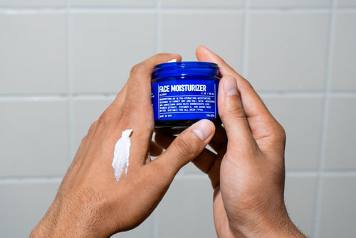 A person holds a blue jar of Blu Atlas Hydrating Face Moisturizer with a dab of white cream on the back of their other hand, shown against a tiled background.