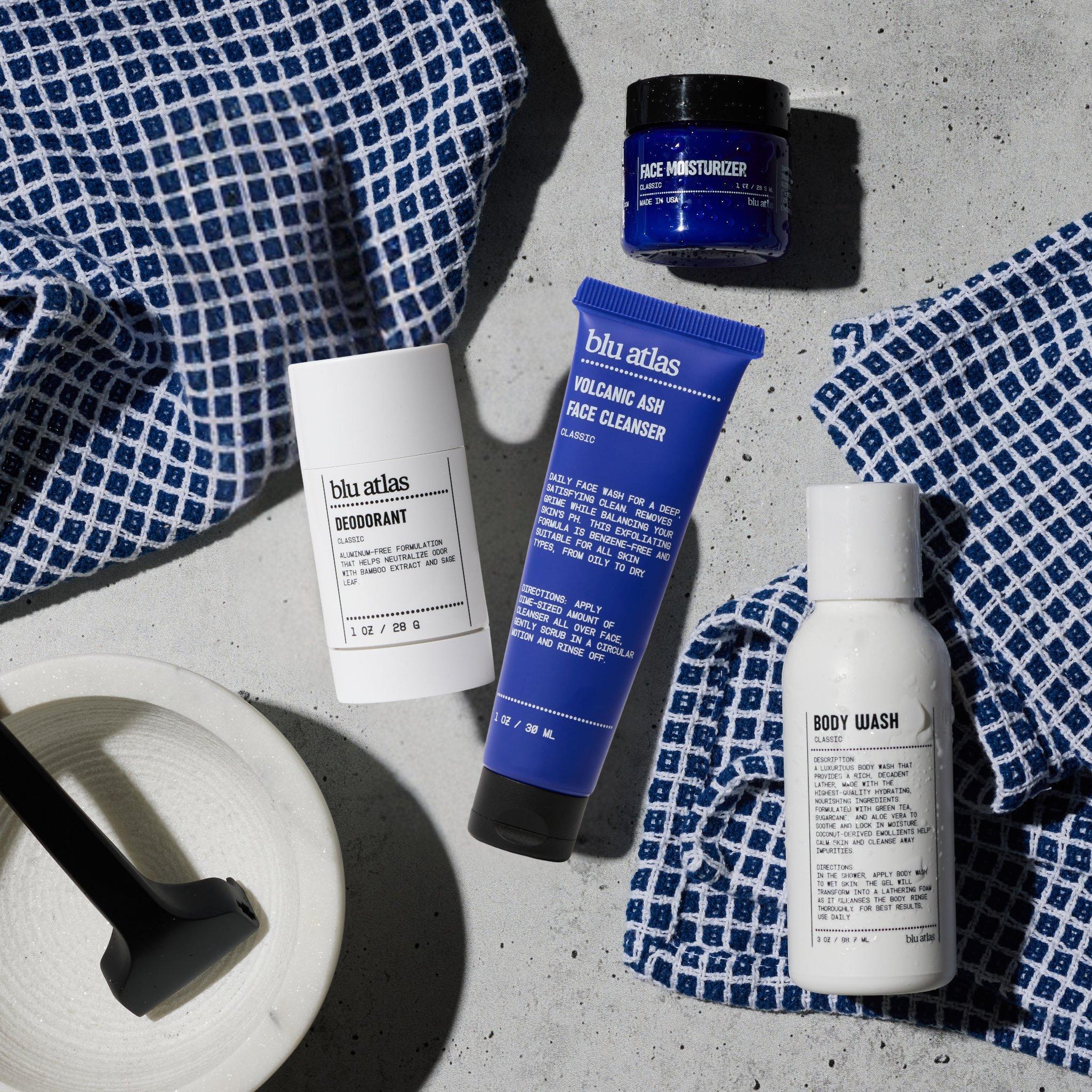 The Blu Atlas Bestseller Mini Bundle—including an aluminum-free men’s deodorant with Bamboo Extract, face cleanser, moisturizer, and body wash—is displayed on a textured surface with blue and white towels and a small brush.