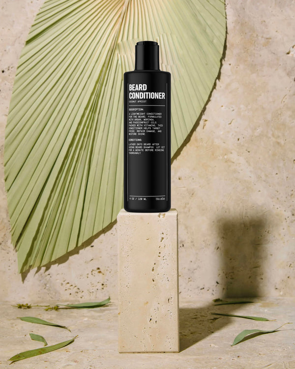 A bottle of Blu Atlas Beard Conditioner, enriched with argan oil, sits elegantly on a beige stone pedestal, framed by a large green palm leaf in the background, promising to repair damage with every use.