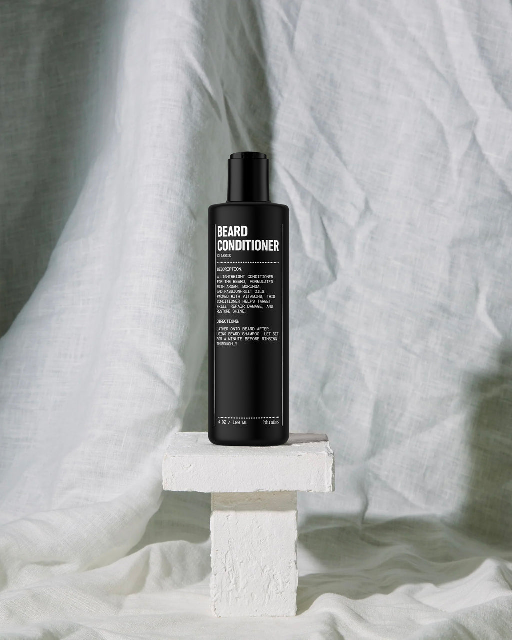 A sleek black bottle of Blu Atlas Beard Conditioner with Argan Oil sits on a white pedestal against a crumpled white fabric backdrop, ready to repair damage and nourish your facial hair.