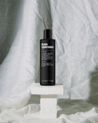 A sleek black bottle of Blu Atlas Beard Conditioner with Argan Oil sits on a white pedestal against a crumpled white fabric backdrop, ready to repair damage and nourish your facial hair.