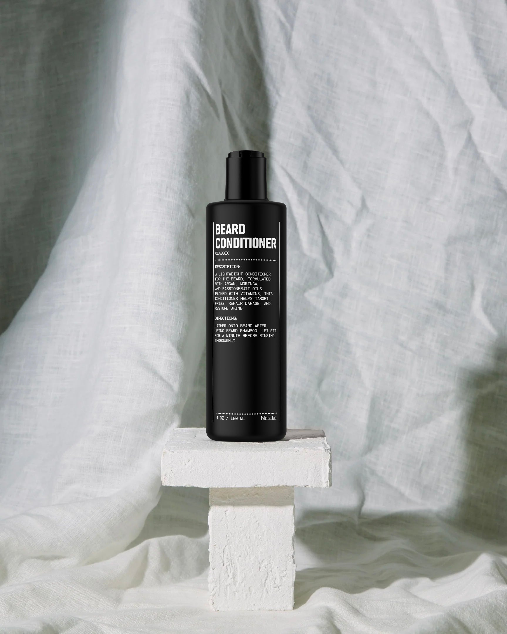 A sleek black bottle of Blu Atlas Beard Conditioner with Argan Oil sits on a white pedestal against a crumpled white fabric backdrop, ready to repair damage and nourish your facial hair.