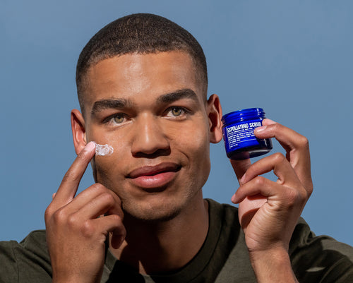 A man applies Blu Atlas Exfoliating Scrub to his cheek while holding a blue jar labeled "Exfoliating Scrub" against a plain blue background.