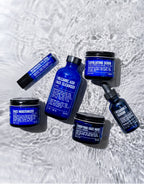An eye-catching Advanced Skincare Set from Blu Atlas shimmers on a reflective surface, showcasing a Volcanic Ash Face Cleanser, an invigorating scrub, and a Vitamin C Serum. Accompanied by a face mask, moisturizer, and lip balm—all elegantly packaged in blue—this collection promises radiant skin.