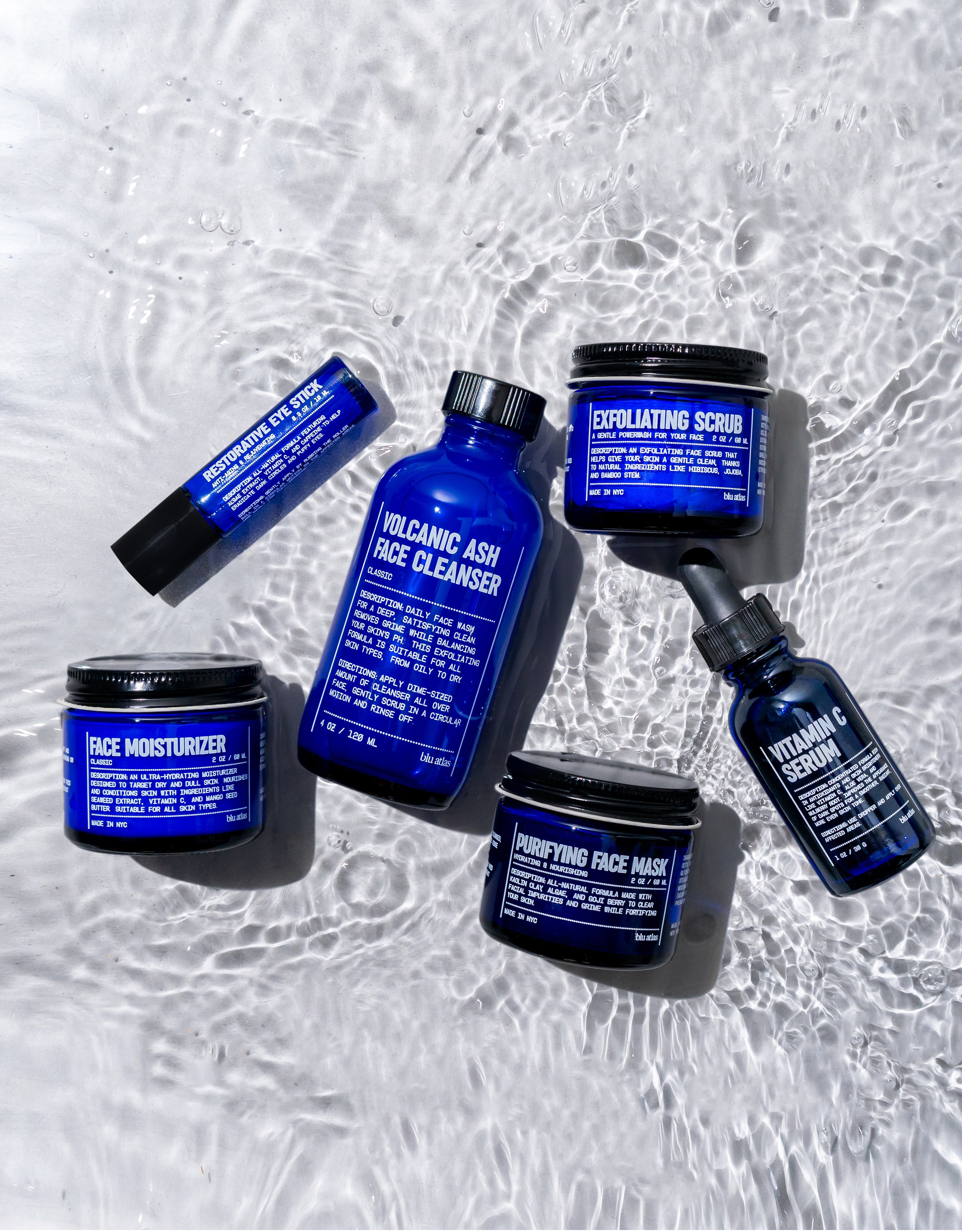 An eye-catching Advanced Skincare Set from Blu Atlas shimmers on a reflective surface, showcasing a Volcanic Ash Face Cleanser, an invigorating scrub, and a Vitamin C Serum. Accompanied by a face mask, moisturizer, and lip balm—all elegantly packaged in blue—this collection promises radiant skin.
