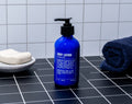 A blue bottle of Blu Atlas Natural Body Lotion with shea butter and a pump sits on a black tiled counter next to a bar of soap and a folded navy towel.