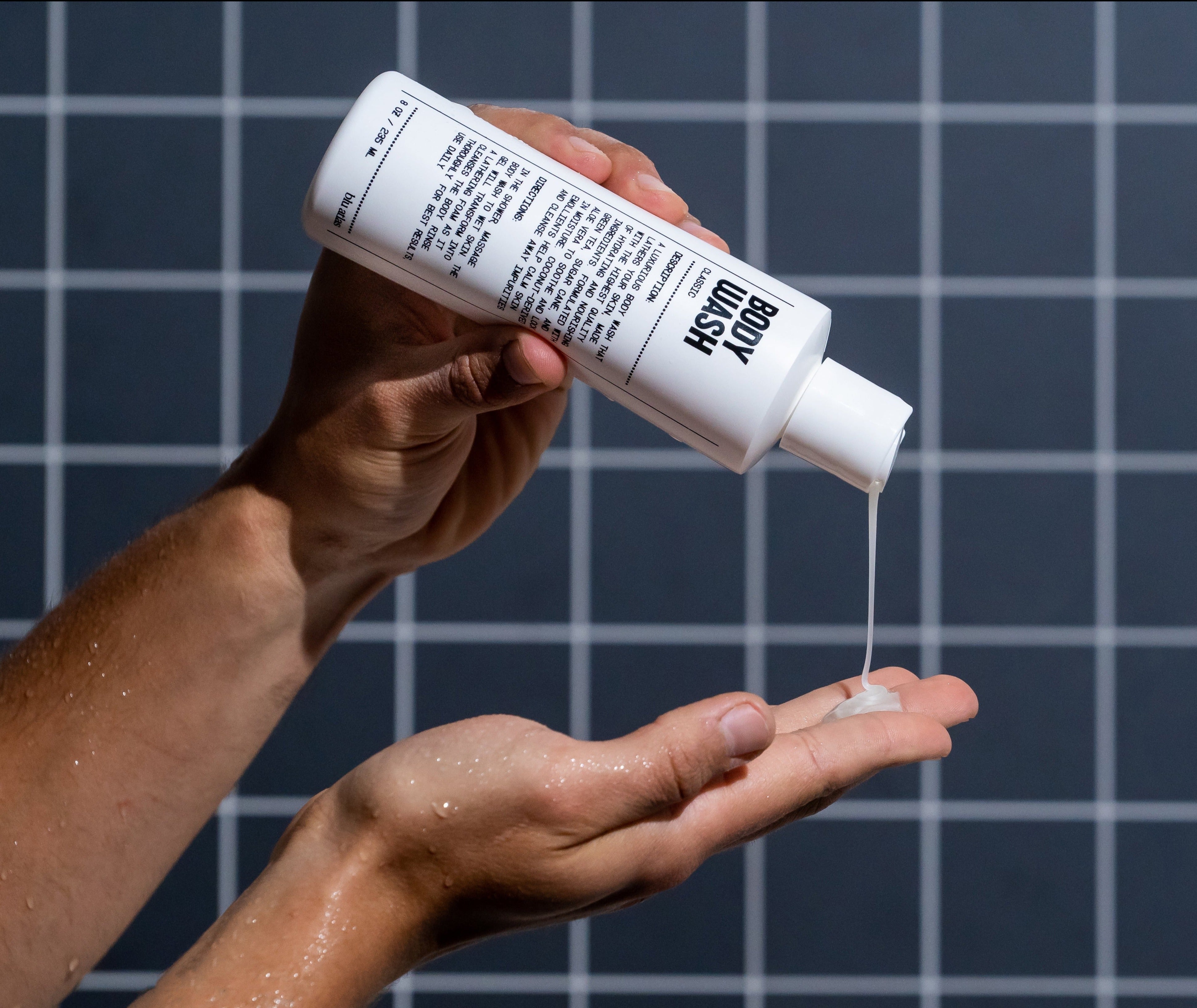 A person pours Blu Atlas Hydrating Body Wash, enriching their hand with hydrating ingredients. The sleek white bottle with black text contrasts against the tiled wall in the background.