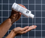 A person pours Blu Atlas Hydrating Body Wash, enriching their hand with hydrating ingredients. The sleek white bottle with black text contrasts against the tiled wall in the background.