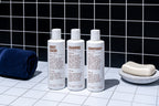The Blu Atlas Shower Refresh Set—three white bottles labeled body wash, shampoo, and conditioner—sits on a tiled surface with a navy towel and soap dish. A black tiled wall provides the backdrop. All products feature natural ingredients.