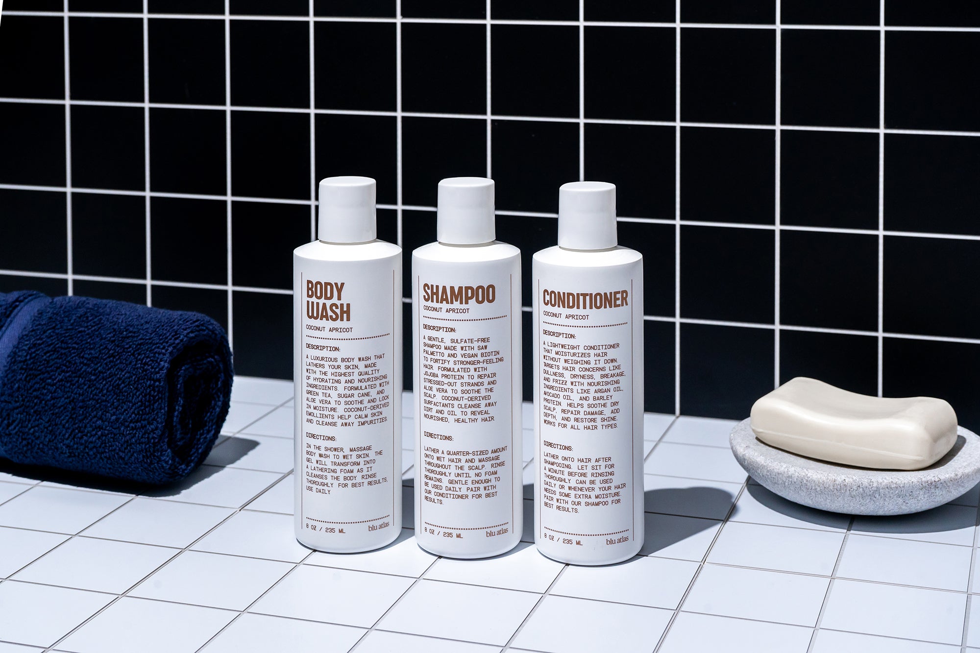 The Blu Atlas Shower Refresh Set—three white bottles labeled body wash, shampoo, and conditioner—sits on a tiled surface with a navy towel and soap dish. A black tiled wall provides the backdrop. All products feature natural ingredients.