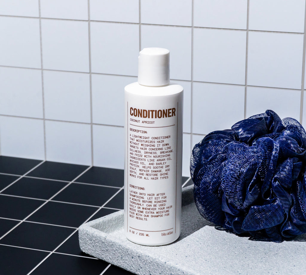 A bottle of Blu Atlas Soothing and Smoothing Natural Conditioner is positioned on a tray next to a dark blue shower puff, all set against a backdrop of white tiles, featuring nourishing ingredients designed to restore shine.
