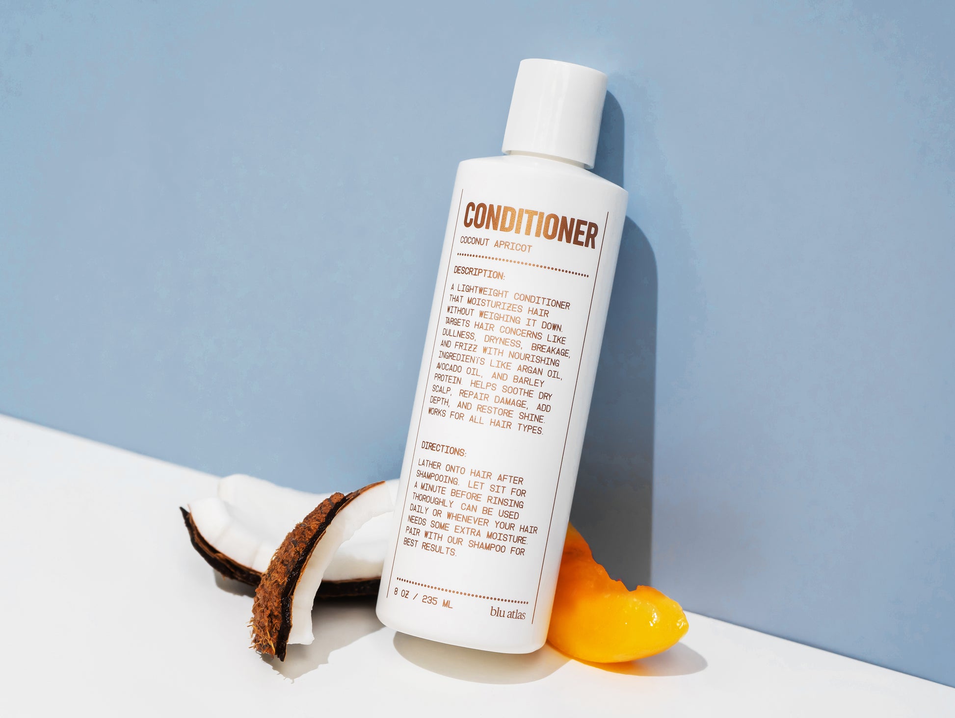 The bottle of Soothing and Smoothing Natural Conditioner by Blu Atlas is positioned against a blue background, accompanied by pieces of coconut shell and an apricot slice, highlighting its nourishing ingredients that are formulated to restore shine.