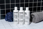 The Blu Atlas Shower Refresh Set—three white bottles labeled conditioner, shampoo, and body wash—is arranged on a tiled surface with a blue shower pouf and rolled gray towel, set against a black tiled wall. Ideal for mens grooming.
