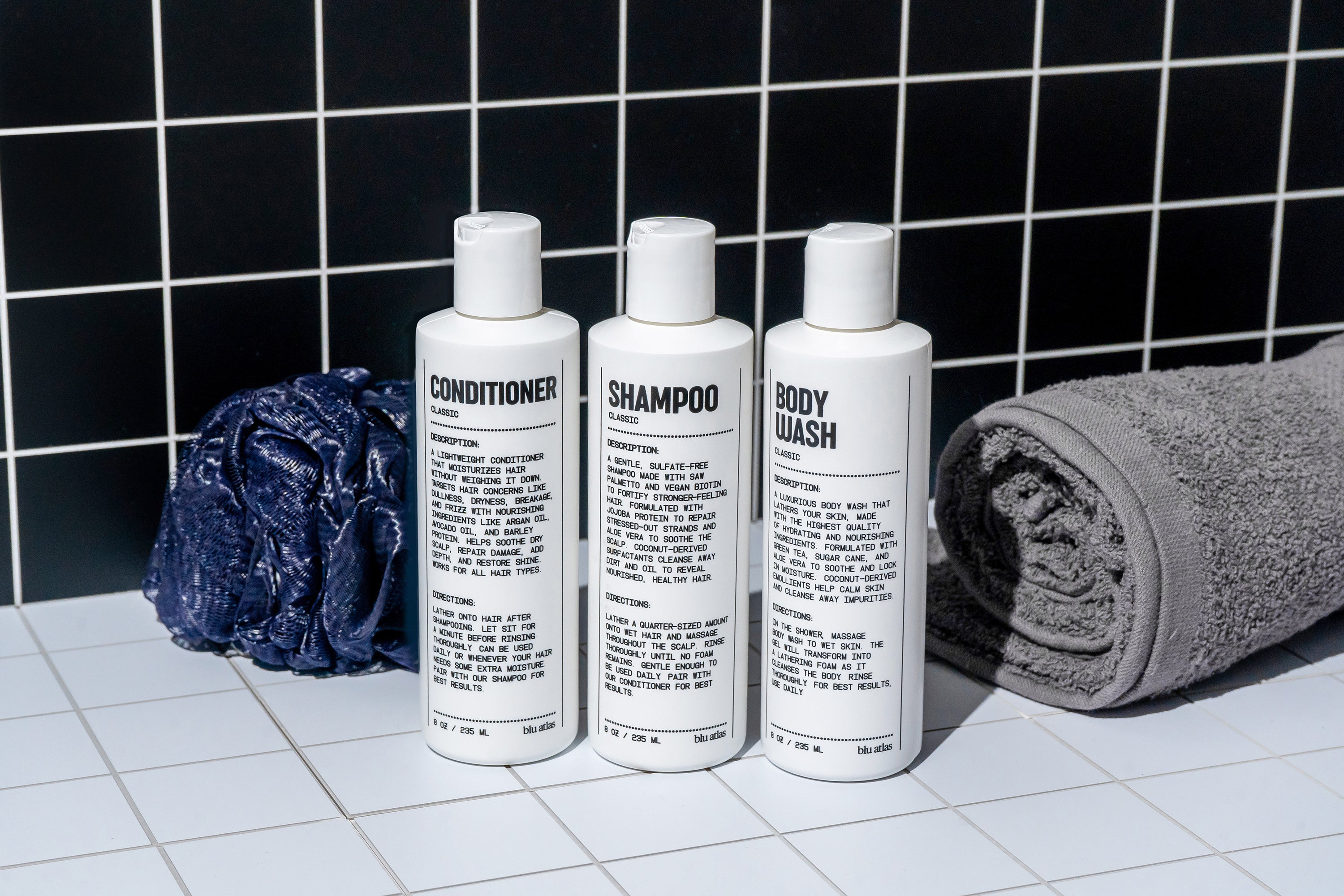 The Blu Atlas Shower Refresh Set—three white bottles labeled conditioner, shampoo, and body wash—is arranged on a tiled surface with a blue shower pouf and rolled gray towel, set against a black tiled wall. Ideal for mens grooming.