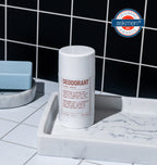 A Blu Atlas Add 48-Hour Natural Deodorant stick, aluminum-free and labeled Coconut & Apricot, sits on a marble tray with a blue soap bar. Black tiled walls in the background highlight its premium quality, enriched with natural Bamboo Extract, and adorned with a Grooming Awards 2022 label.