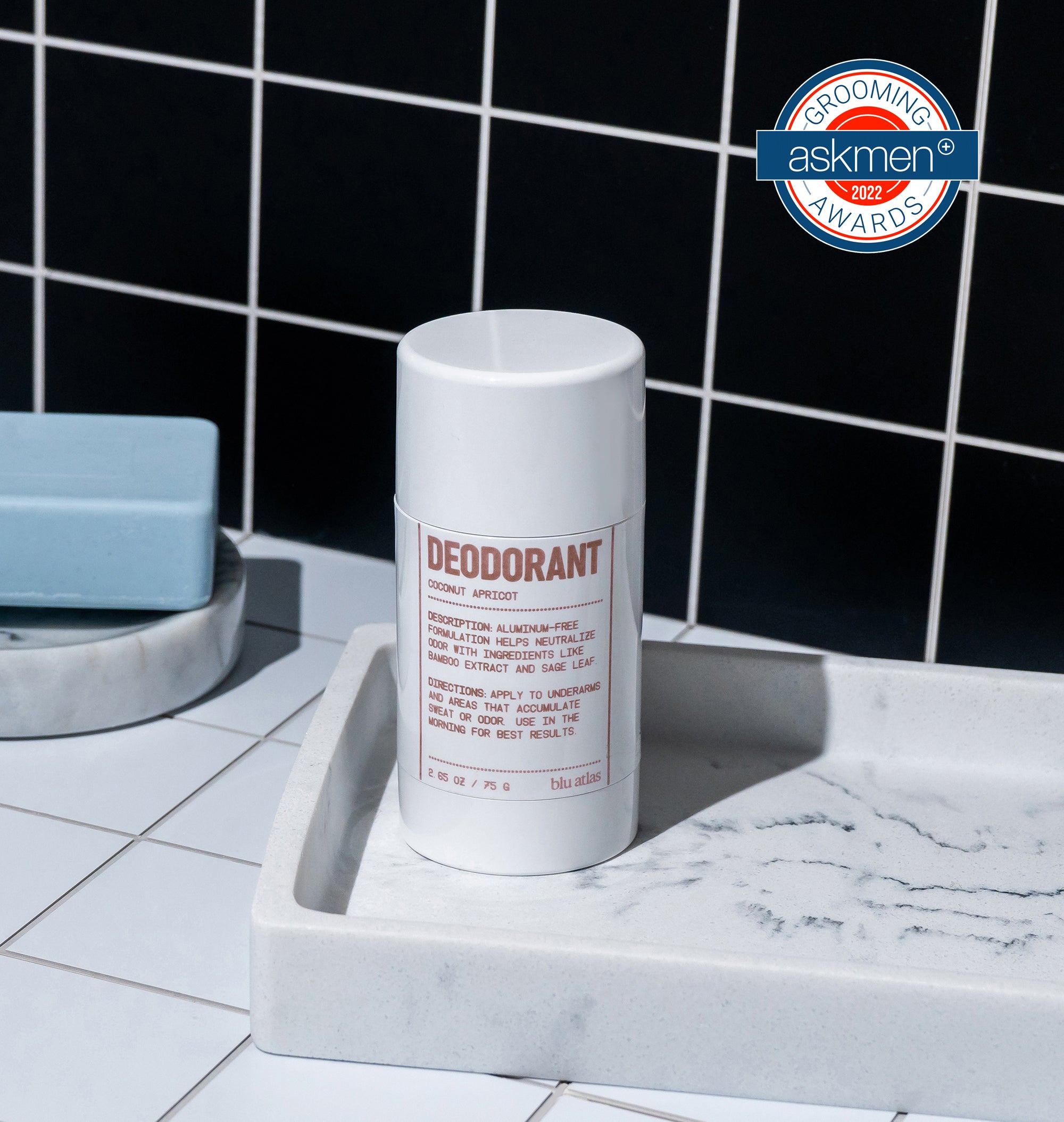 A Blu Atlas Add 48-Hour Natural Deodorant stick, aluminum-free and labeled Coconut & Apricot, sits on a marble tray with a blue soap bar. Black tiled walls in the background highlight its premium quality, enriched with natural Bamboo Extract, and adorned with a Grooming Awards 2022 label.