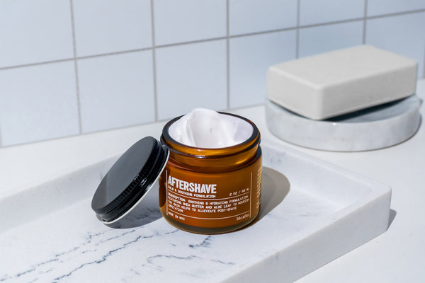 An open amber jar of Blu Atlas Natural Aftershave rests on a white tray beside a closed soap dish in a tiled bathroom, its soothing formula enriched with rose water to gently care for sensitive skin.
