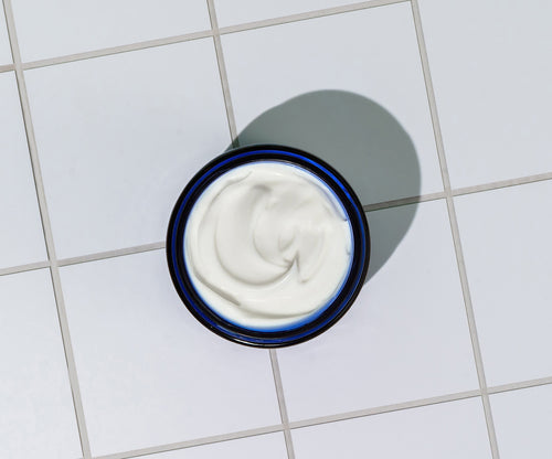 A jar of Blu Atlas Hydrating Face Moisturizer, with its creamy white formula, rests on a white tiled surface, casting a shadow to the right.