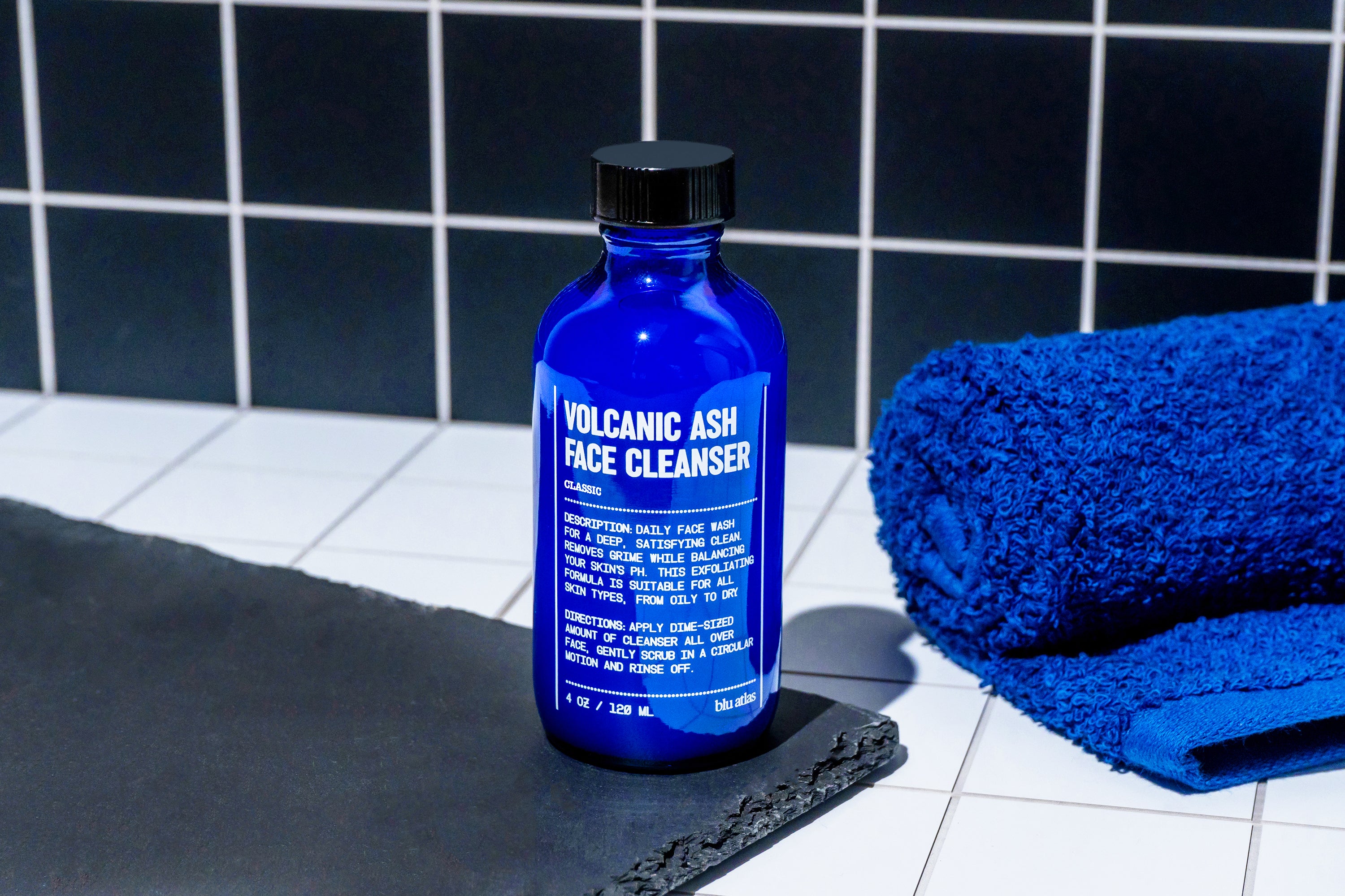 A blue bottle of Blu Atlas Volcanic Ash Gel Cleanser, suitable for all skin types, stands on a tiled surface beside a folded blue towel, with a black tile wall behind it.