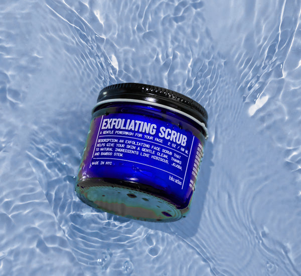 A jar of Blu Atlass Gentle Exfoliating Scrub for All Skin Types, with a blue label and natural exfoliants, floats on rippling water, making it essential for your grooming routine.