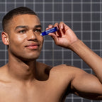 A man stands before a tiled wall, using the Blu Atlas Men's Restorative Eye Stick For Puffy Eyes from a blue container to address stubborn dark circles and achieve a rejuvenated appearance.